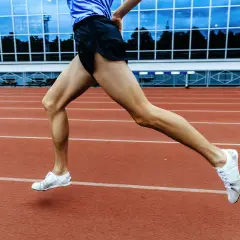 Sylwetka biegacza w ruchu na bieżni stadionu. Mięśnie nóg naprężone, białe buty sportowe unoszą się nad czerwonym torem.