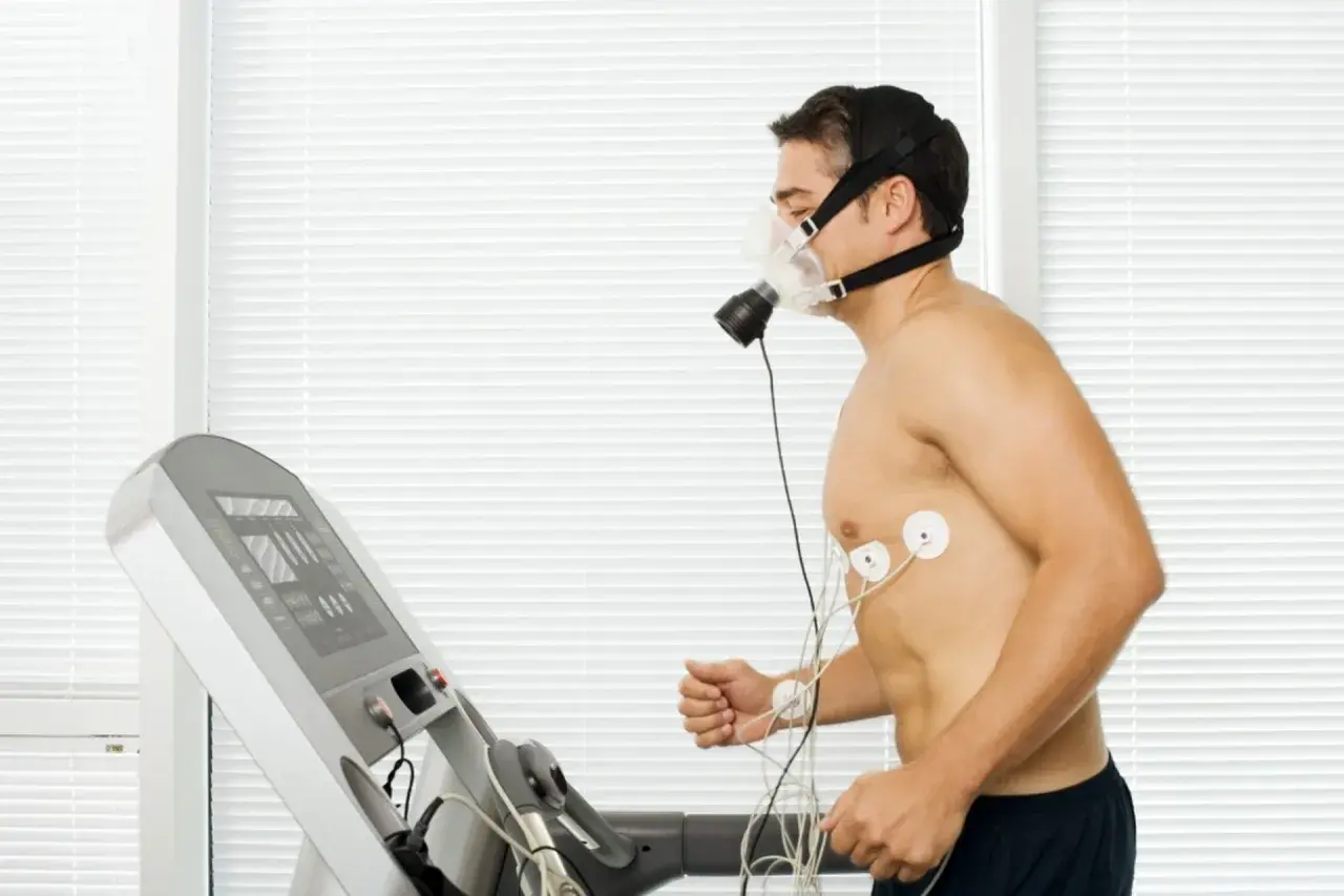 Mężczyzna w masce biegający na bieżni, podłączony do aparatury EKG. Wyniki testu VO2max w tabeli na ekranie bieżni.