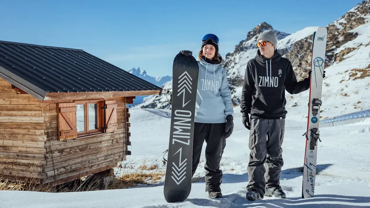 Para na tle g&oacute;rskiej chatki prezentuje stylowy ubi&oacute;r na narty i snowboard. Ona w jasnej bluzie, on w ciemnej.