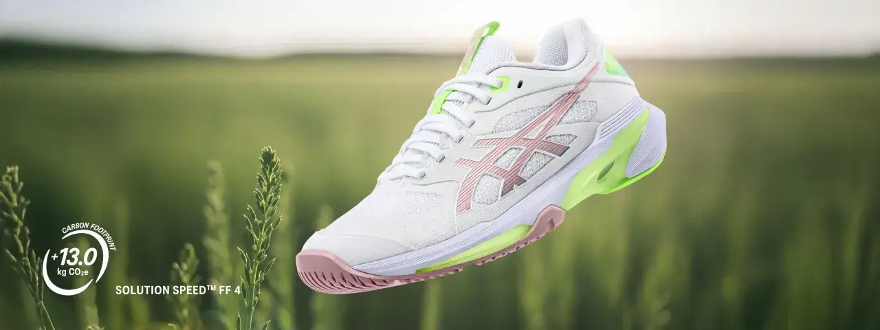 Białe buty tenisowe z r&oacute;żowymi i zielonymi akcentami od firmy Asics, model Solution Speed FF 4, unoszą się na tle zielonego pola.